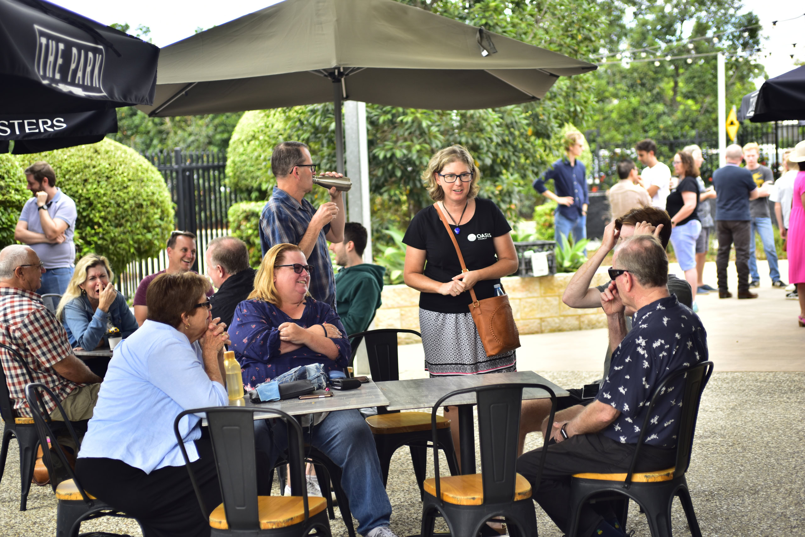 People gathered at an outdoor café under umbrellas, engaged in conversation. A woman wearing an ‘Oasis’ shirt stands talking to a seated group, with others socialising in the background. | Easter at Oasis Church