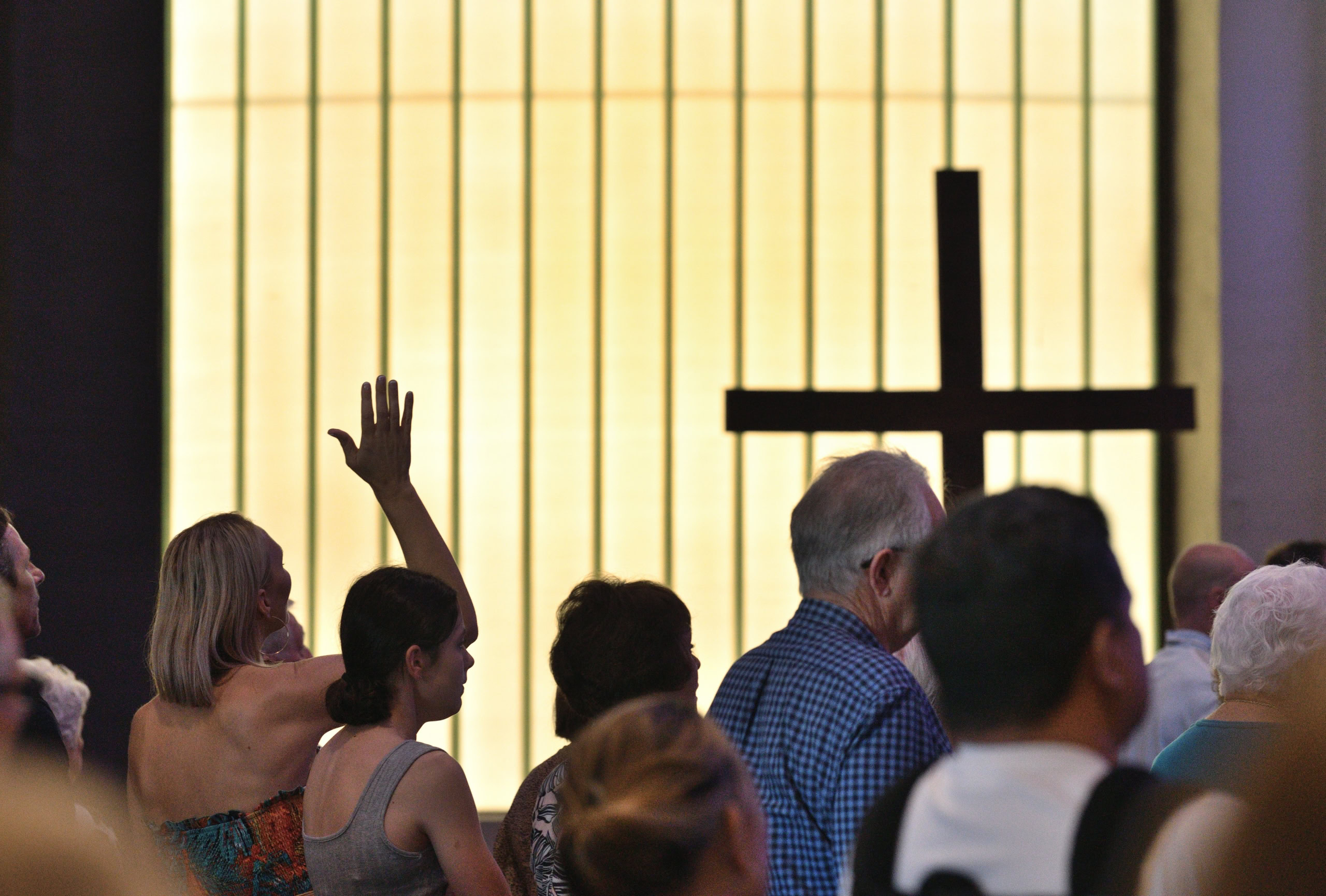A congregation gathers in worship, with a silhouetted cross in front of illuminated windows. A woman raises her hand in praise | Easter at Oasis Church