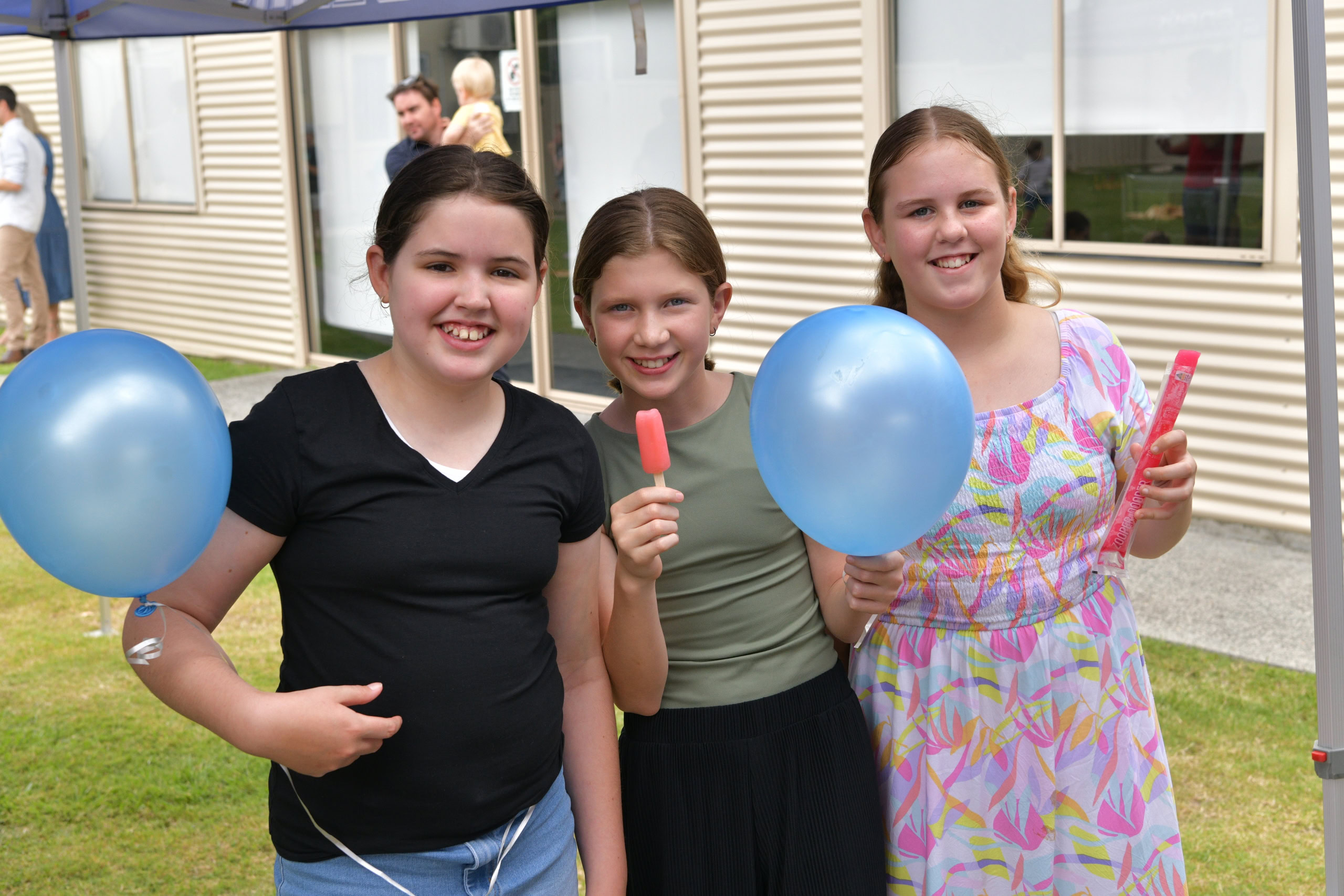 Three smiling girls stand under a tent, holding blue balloons and frozen treats, enjoying a community event | Easter at Oasis Church