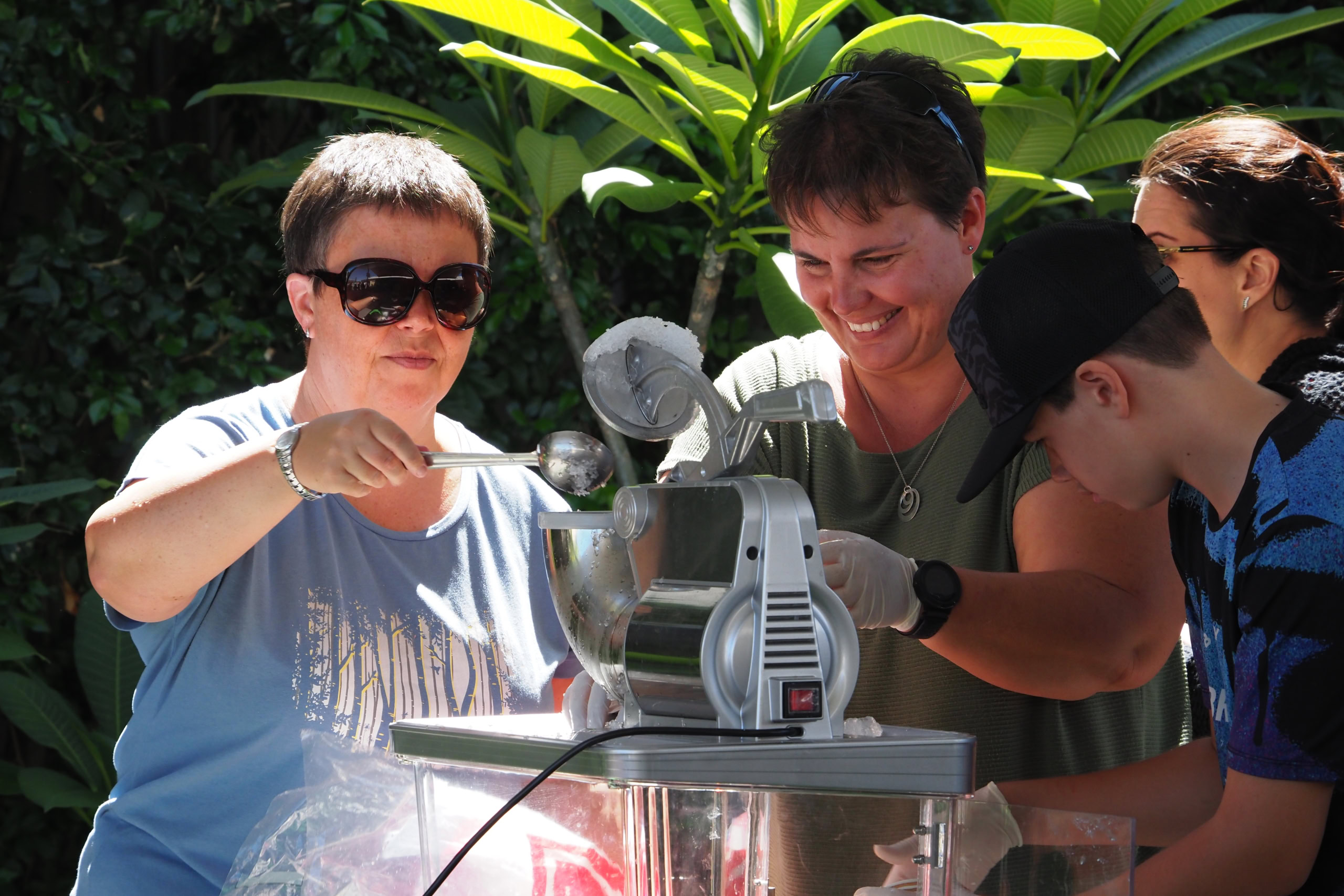 Two women smile while preparing snow cones at an outdoor event, using a shaved ice machine as a young boy watches | Easter at Oasis Church