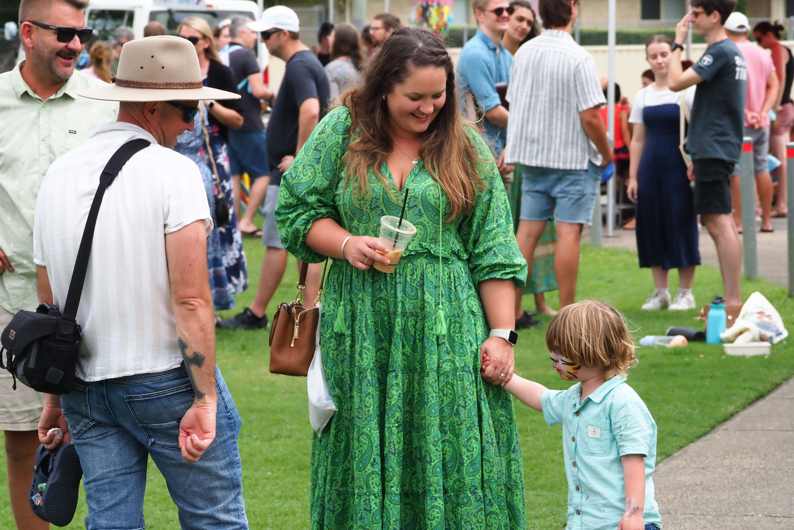 A woman in a green dress holds a drink while walking with a young child with face paint. People are mingling in the background.