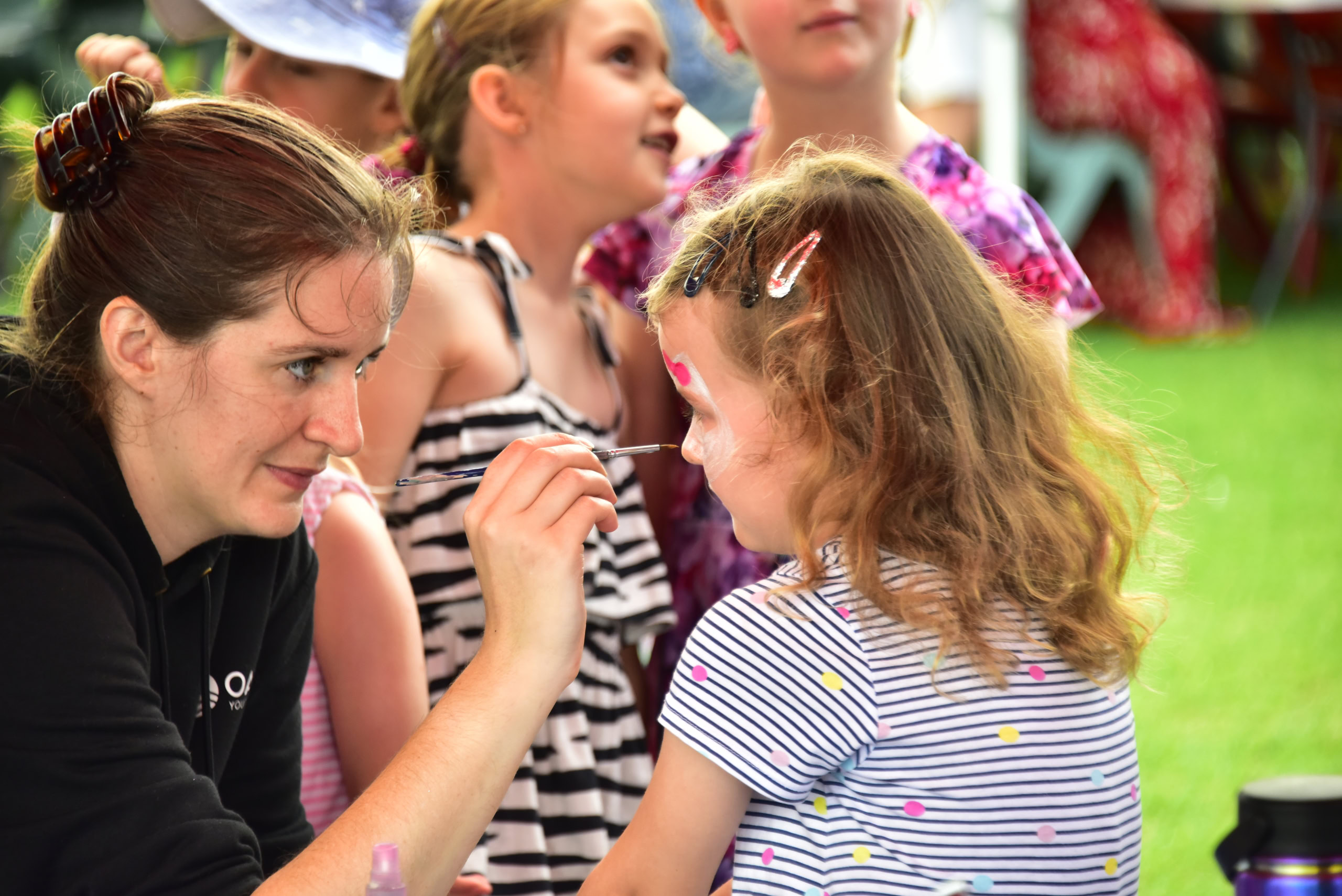 A young girl with curly hair sits patiently as a woman applies face paint to her cheek. Other children in colourful outfits watch in the background | Easter at Oasis Church