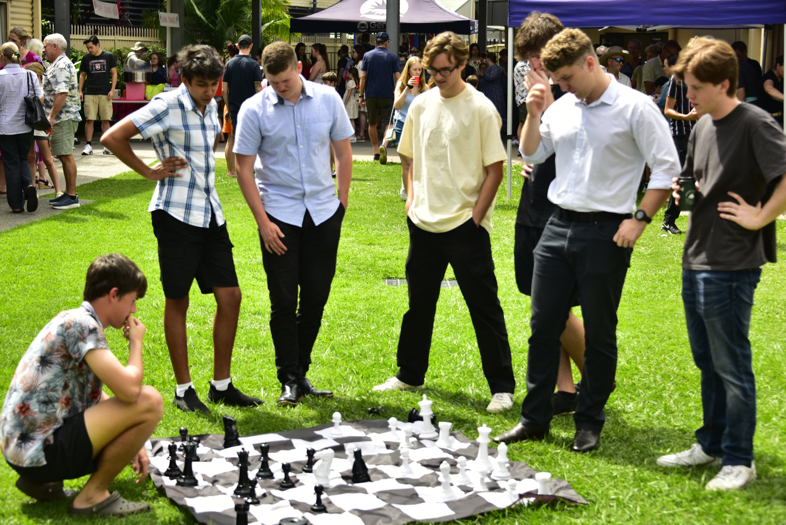 A group of young men stands around a large outdoor chess set, watching a player crouched down to make a move. Market stalls and event tents are visible in the background | Easter at Oasis Church