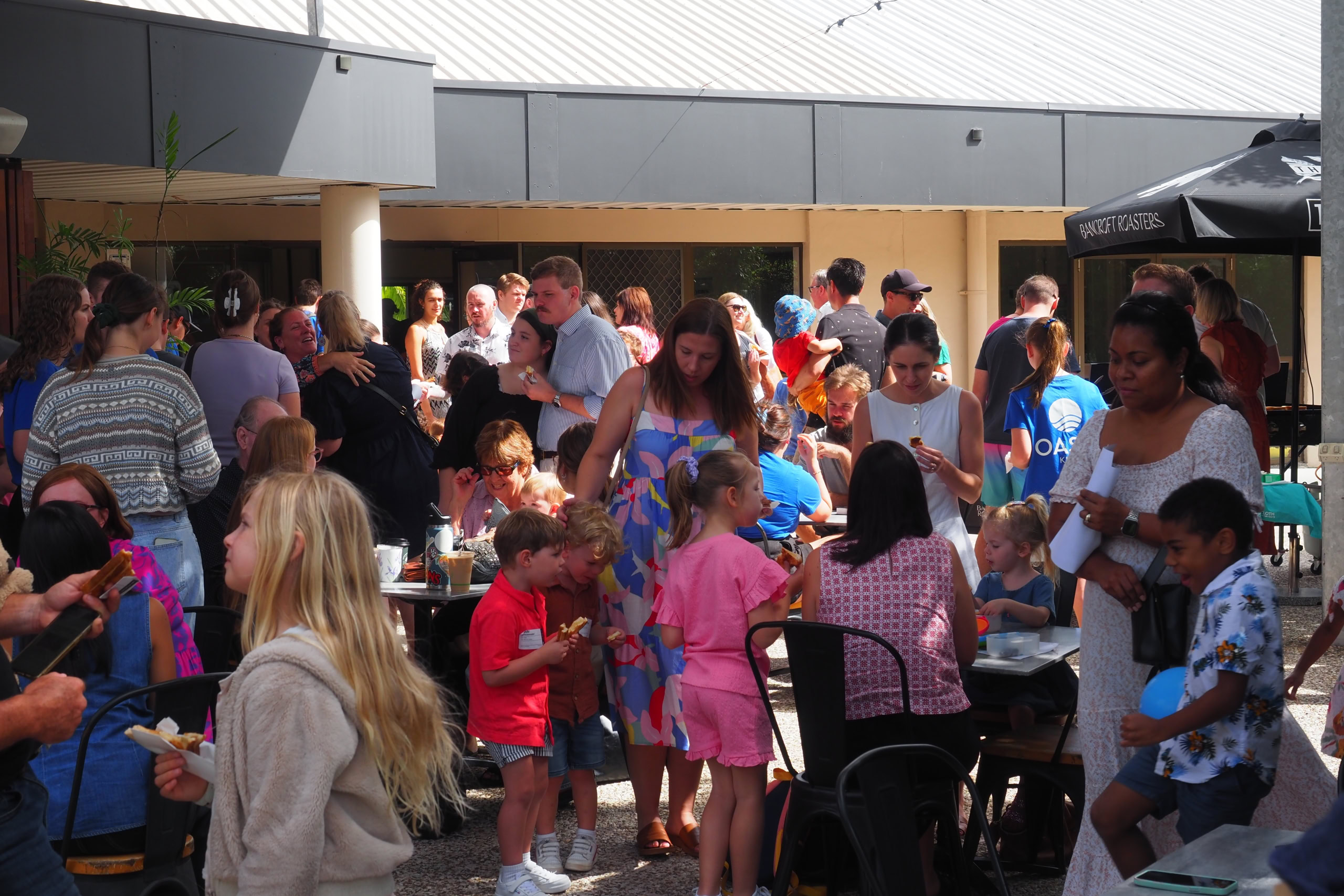 A busy courtyard filled with people of all ages, chatting, eating, and enjoying a lively event under tents.