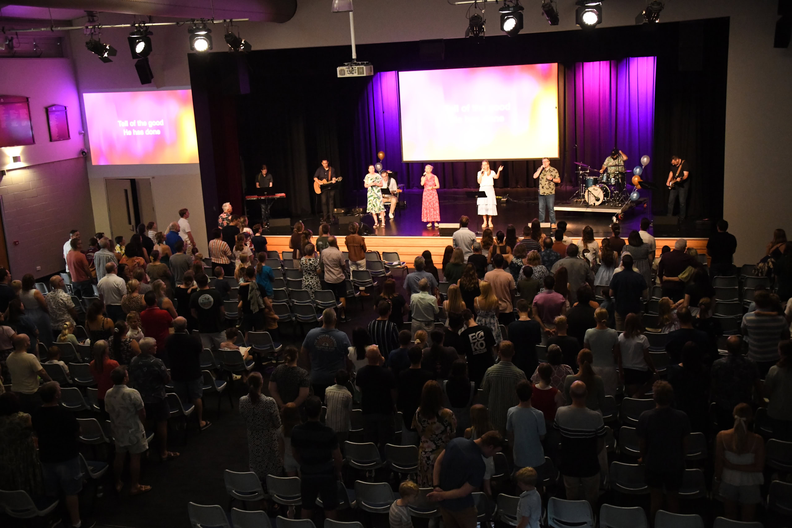 A band leads worship on stage with singers, musicians, and a vibrant purple-lit backdrop. The congregation stands and sings along.