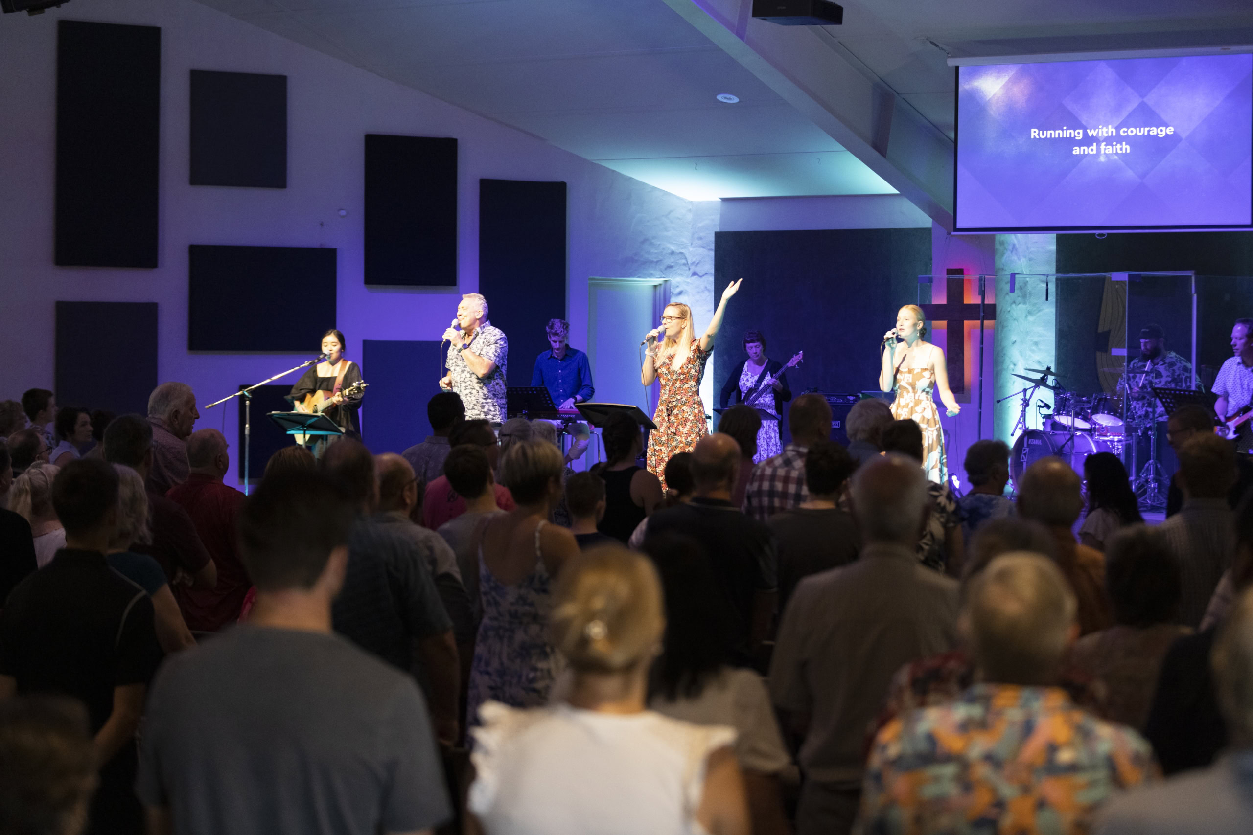 A worship band leads a church congregation in singing, with singers and musicians on stage under blue lighting. A screen displays the lyrics ‘Running with courage and faith,’ and a cross is illuminated in the background.